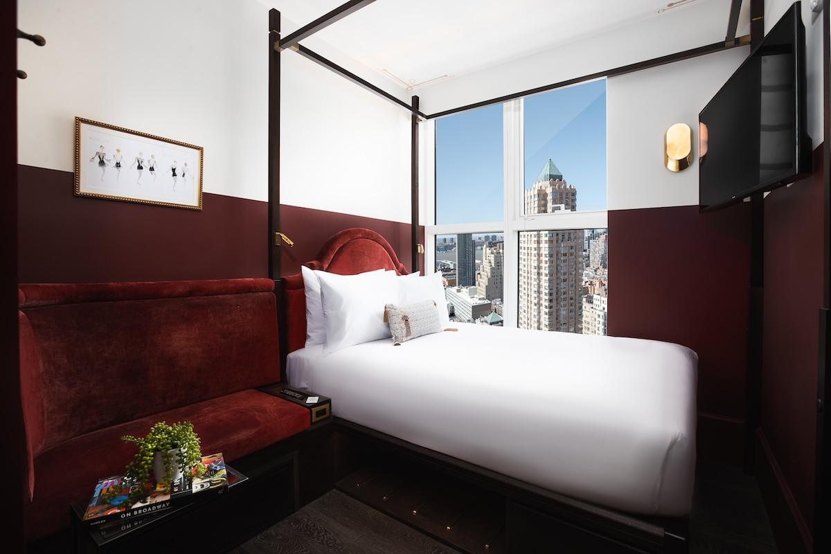 A Cozy Queen room: a four-poster queen bed, a red velvet banquette and a tall window onto Midtown towers